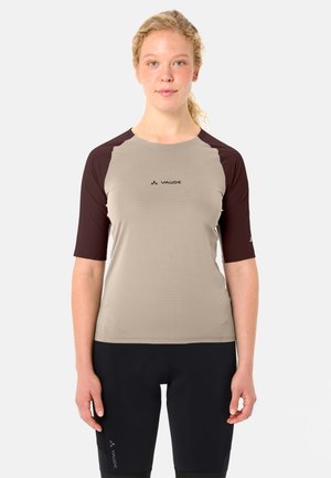 Giovane donna con capelli ricci chiari indossa una maglia da ciclismo beige e marrone scuro e pantaloncini da ciclismo imbottiti neri su sfondo bianco.