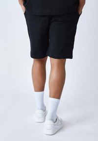Un short noir avec une texture lisse, deux poches latérales, associé à des baskets blanches et des chaussettes blanches côtelées, vu de dos.