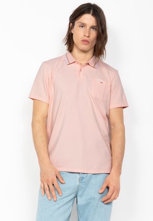 Jeune homme aux cheveux mi-longs portant un polo rose à manches courtes et un jean bleu clair, debout devant un fond blanc.