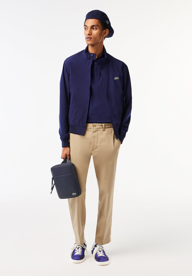 Homme portant une veste bleu marine, un béret assorti, un pantalon beige et des baskets bleues, tenant une pochette bleu marine dans une pose debout contre un fond blanc.