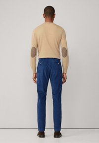 Maglione in wool beige con toppe marroni sui gomiti, abbinato a pantaloni in cotone blu scuro con tasche anteriori e posteriori. Scarpe marroni completano il look.