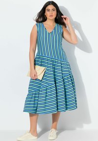 Ärmlös blå klänning med vertikala ränder, V-ringning och flerlagers nederkant. Kombination med en vävd kuvertväska och beige sneakers.