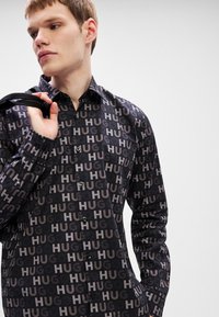 Μαύρο πουκάμισο με κουμπιά, που διαθέτει επαναλαμβανόμενο τύπωμα "HUG" σε διάφορες αποχρώσεις. Κατασκευάζεται από απαλό ύφασμα, με κλασικό γιακά και μακριά μανίκια.