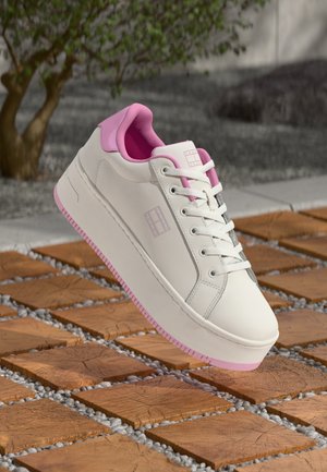 Baskets plateformes blanches avec des accents roses flottant au-dessus d'un sol en carreaux de bois et de galets, avec un arbre flou en arrière-plan.