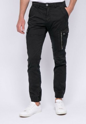 Homme portant un pantalon cargo noir avec une poche latérale zippée et des poignets élastiques, associé à des baskets blanches et une chemise blanche.