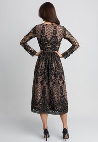 Czarna koronkowa sukienka z długimi rękawami, półprzezroczystą warstwą, skomplikowanymi wzorami kwiatowymi, podkreśloną talią i długością do połowy łydki. Sandały na wysokim obcasie.