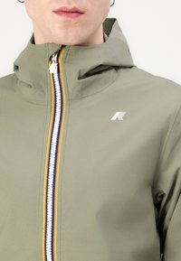 Chaqueta verde oliva con cuello alto, cremallera frontal con rayas multicolores y un logo blanco en el lado izquierdo del pecho. Textura suave.