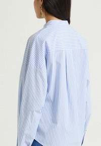 Blusa a righe azzurro chiaro e bianco con una vestibilità rilassata, caratterizzata da una piega sul retro e tessuto morbido. Maniche lunghe con dettaglio polsino.
