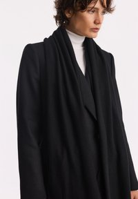 Czarny wełniany płaszcz z drapowanym przodem i dopasowaną sylwetką. Posiada kołnierz typu szal, długie rękawy oraz fakturowany materiał.