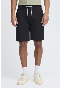 Svarte shorts laget av lett stoff, med snøring i livet, sidelommer og rett snitt, brukt med hvite sneakers og sokker.