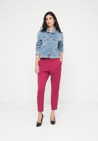 Τζιν μπουφάν με τέσσερις μπροστινές τσέπες, μπλε χρώμα, συνδυασμένο με εφαρμοστά ματζέντα παντελόνια και μαύρα ψηλά τακούνια. Απλό φόντο, όρθια στάση.