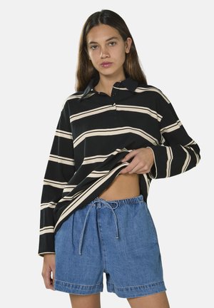 Ung kvinde løfter en sort og beige stribet langærmet skjorte, iført blå denim shorts med snøre, mod en ensfarvet lys baggrund.