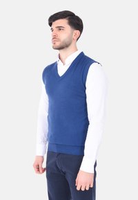 Gilet azzurro lavorato a maglia con scollatura a V, orlo e fori per le braccia a coste, indossato sopra una camicia bianca con colletto, modello posato su uno sfondo neutro.