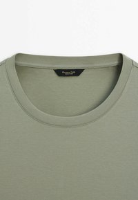 T-shirt vert olive avec un col rond, une texture lisse, et une étiquette avec des lettres dorées, placée à l'arrière du cou.