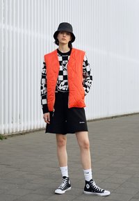 Colete acolchoado vermelho sobre uma camisa de mangas longas com padrão preto e branco, calções pretos, meias brancas e sapatilhas pretas de cano alto.
