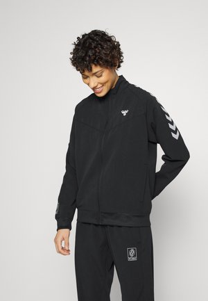 Schwarze Zip-Jacke mit hohem Kragen, aus strukturiertem Stoff, mit reflektierenden Chevron-Akzenten an den Ärmeln und Logo auf der Brust.