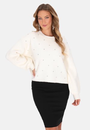 Kvinde med langt bølget hår iført hvid sweater med små pynt og sort nederdel, poserer med den ene hånd på hoften foran en ensfarvet baggrund.