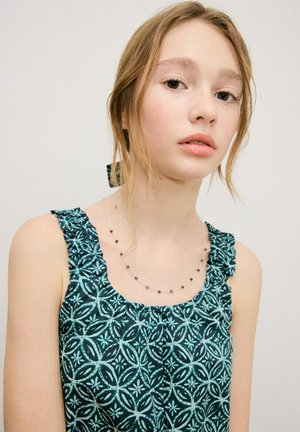 Jeune femme aux cheveux châtains attachés, portant un haut sans manches vert foncé à motifs et un délicat collier de perles, sur un fond uni.