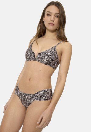 Mujer con cabello largo y castaño, vistiendo un bikini negro y beige con estampado y tirantes delgados, de pie contra un fondo claro y liso.