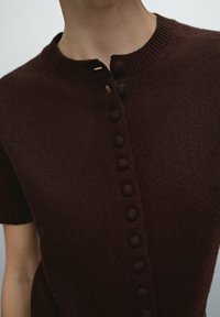 Zbliżenie na osobę noszącą ciemno-brązowy, dzianinowy top z krótkim rękawem, z rzędem zakrytych guzików z przodu i prążkowanym kołnierzykiem.