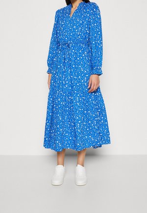 Frau in einem langärmeligen, blauen Kleid mit Blumenmuster und geraffter Taille, dazu weiße Sneaker, steht vor einem schlichten hellen Hintergrund.