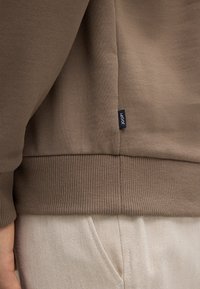 Felpa marrone con polsini e orlo a coste, presenta un piccolo etichetta nera di lato. Texture liscia, design semplice e colore uniforme.