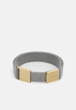 Bracelet en maille argentée avec deux fermoirs rectangulaires dorés gravés "GUESS" sur fond blanc.
