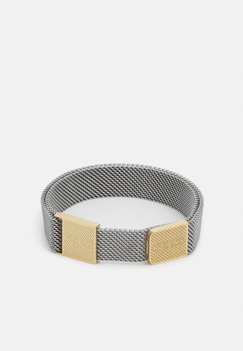 Bracelet en maille argentée avec deux fermoirs rectangulaires dorés gravés "GUESS" sur fond blanc.