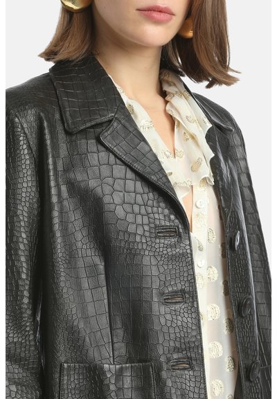 Vrouw in een zwart leren jasje met krokodillenpatroon over een doorschijnende witte blouse met gouden cirkelontwerpen en een ruffelcollar.