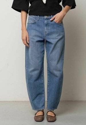 Persona che indossa jeans azzurri larghi, top nero e scarpe basse intrecciate marroni, in piedi davanti a uno sfondo chiaro uniforme.