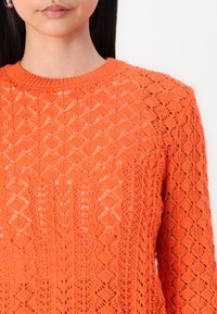 Maglione arancione lavorato a maglia con maniche lunghe, caratterizzato da un motivo a rombi e una trama a tessitura aperta. Collo rotondo con bordi a coste.