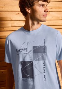 T-shirt en coton bleu clair présentant des motifs graphiques abstraits noirs, comprenant du texte et des formes, sur un fond en bois.