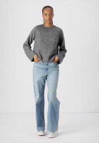 Pull en tricot gris avec des manches larges, associé à un jean évasé bleu clair. Le mannequin porte des baskets blanches et se tient devant un fond uni.