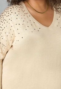 Woman wearing a beige V-neck sweater with scattered shiny sequins on the shoulder and a gold chain necklace.