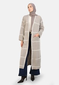 Cardigan long à ouverture frontale en beige avec un motif à carreaux et des accents blancs. Comprend deux poches avant et des manches larges. Tissu tricoté texturé.