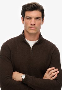 Bruine gebreide trui met een hoge kraag en ritsdetail, gedragen over een wit shirt. Model heeft de armen gekruist en draagt een zilveren horloge.