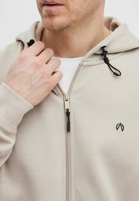 Haut à capuche zippé beige avec une fermeture éclair à l'avant, un cordon de serrage à la capuche et un petit logo sur la poitrine. Présente une texture lisse et des manches ajustées.