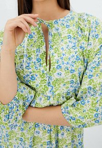 Frau passt den Bindegürtel eines langärmligen Kleides in Grün- und Blaublütenmuster mit elastischen Bündchen und gerafftem Taillenbund an.