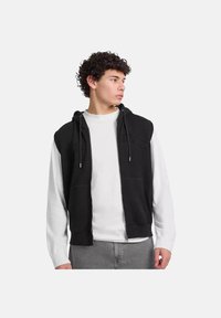 Sudadera sin mangas negra con cremallera frontal, cordones para la capucha y dos bolsillos frontales. Lleva puesta sobre una camiseta de manga larga blanca.