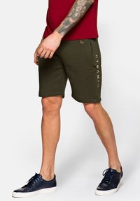 Oliwkowe bawełniane szorty o luźnym kroju, z podwiniętym dołem i złotym nadrukiem "DYNAMIC" z boku. Noszone z ciemnymi sneakersami.