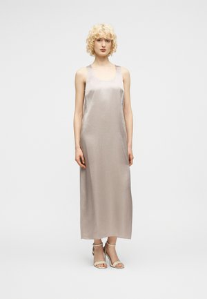 Kvinde med krøllet blondt hår iført en ærmeløs sølvfarvet satin maxikjole og lyse ankelseler med hæl, stående mod en ensfarvet baggrund.