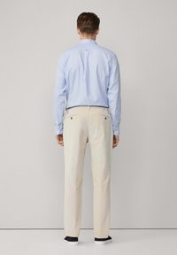 Camisa de botones azul claro y pantalones beige con bolsillos traseros, de pie sobre un fondo liso. La camisa tiene mangas largas y un cuello.