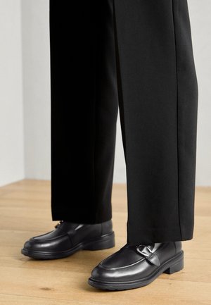 Mocasines de cuero negro con punta redonda, tira decorativa y tacón bajo, combinados con pantalones negros, sobre un suelo de madera.