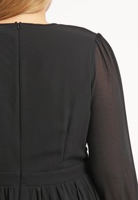 Czarna sukienka z długimi przezroczystymi rękawami, okrągłym dekoltem i dopasowanym topem z marszczoną talią. Zapięcie na zamek z boku na plecach.