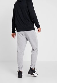 Uomo che indossa pantaloni della tuta grigi chiari, felpa nera e sneaker bianco e nere in piedi davanti a uno sfondo bianco semplice.