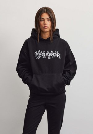 Ung kvinde med langt mørkt hår iført en sort hættetrøje med "PEGADOR" og blomsterdesign, stående mod en ensfarvet lys baggrund.