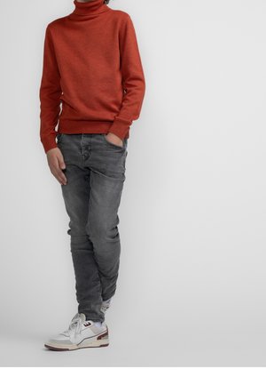 Persona con suéter de cuello alto color óxido, jeans grises y zapatillas blancas, de pie con una mano en el bolsillo frente a un fondo gris liso.