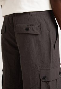 Pantalon cargo gris foncé en tissu léger, présentant plusieurs poches et des boutons sur les côtés, avec une texture froissée.