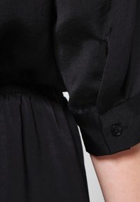 Vestido negro con una textura suave y lisa, cintura fruncida y mangas remangadas sujetas con botones negros.
