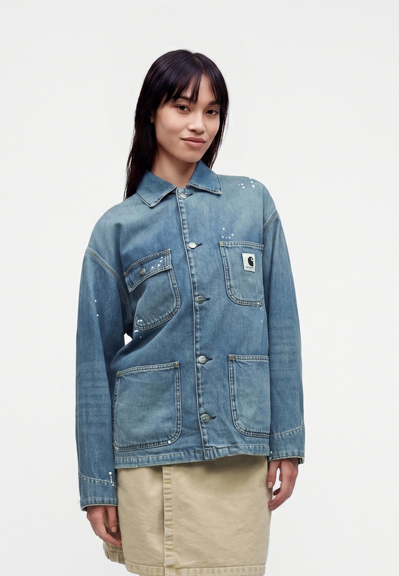 Νεαρή γυναίκα φοράει μπλε denim μπουφάν με κουμπιά και μεγάλες τσέπες, συνδυασμένο με μια μπεζ φούστα σε απλό λευκό φόντο.